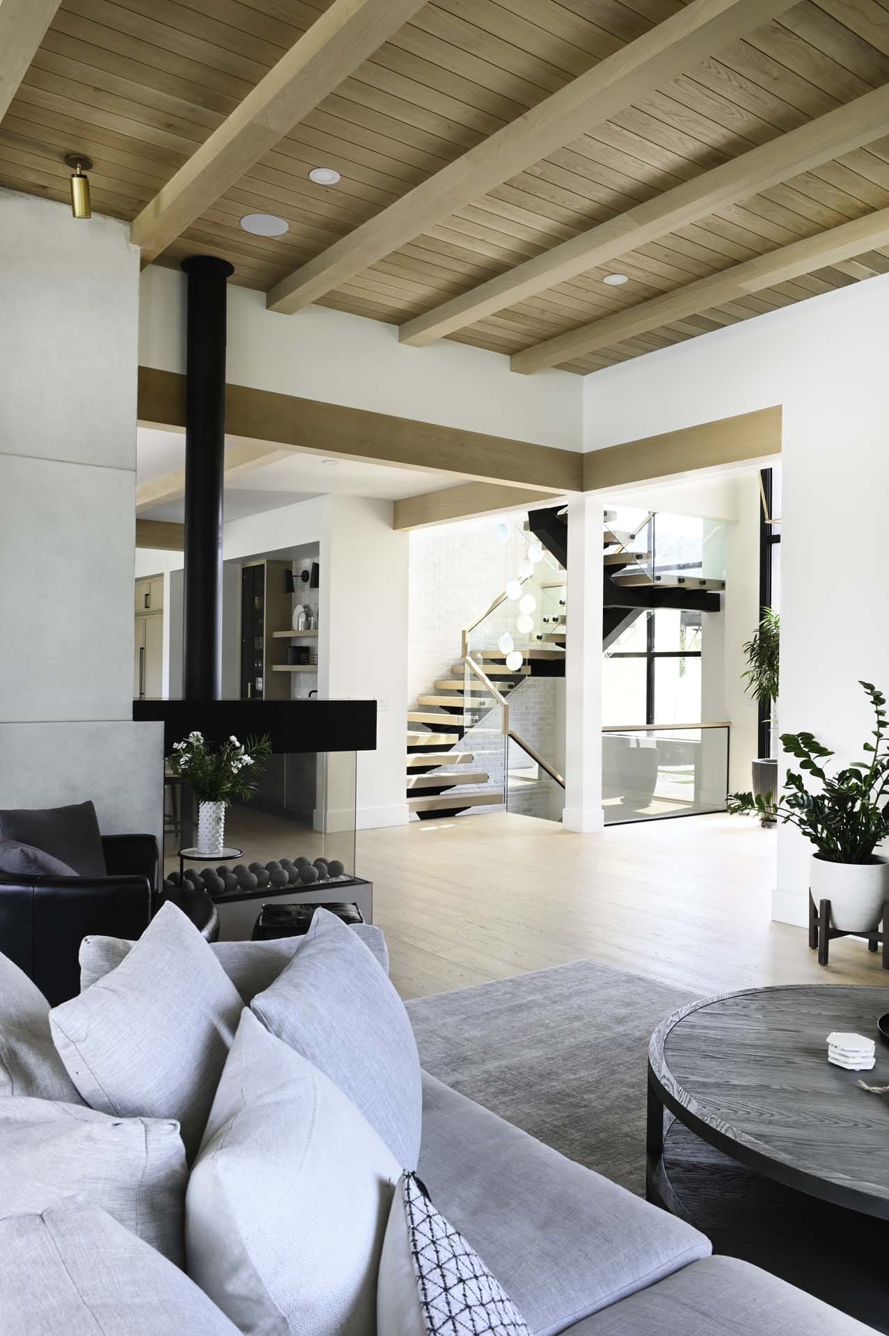 contemporary_home_interior (12) A modern living room showcases the expertise of home designers, featuring a light wood ceiling, gray sofa, and round coffee table. The view reveals a staircase with a glass railing, while large windows flood the space with natural light.