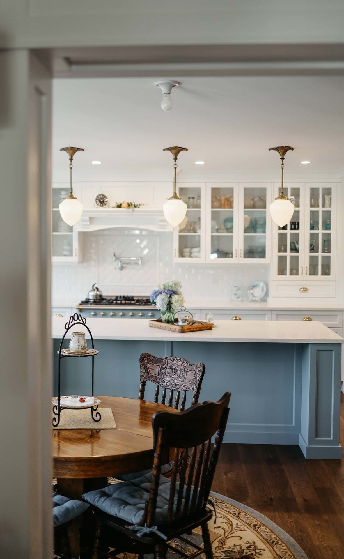 A kitchen designed with white cabinets, a blue island, and elegant pendant lights showcases the artistry of home designers. The wooden dining table with chairs complements the floral centerpiece on the island, reflecting the essence of custom home design.