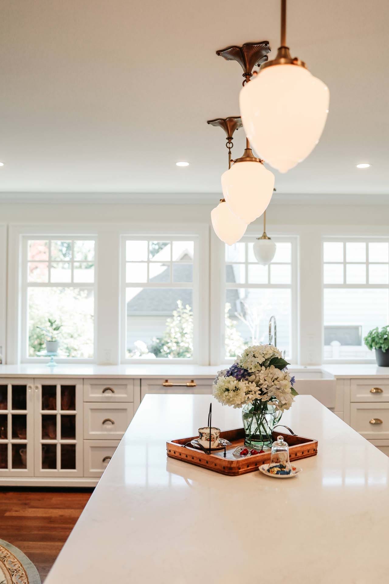 A bright kitchen showcases a custom home design with a long white countertop, pendant lights, large windows, and a tray adorned with flowers and serving items.