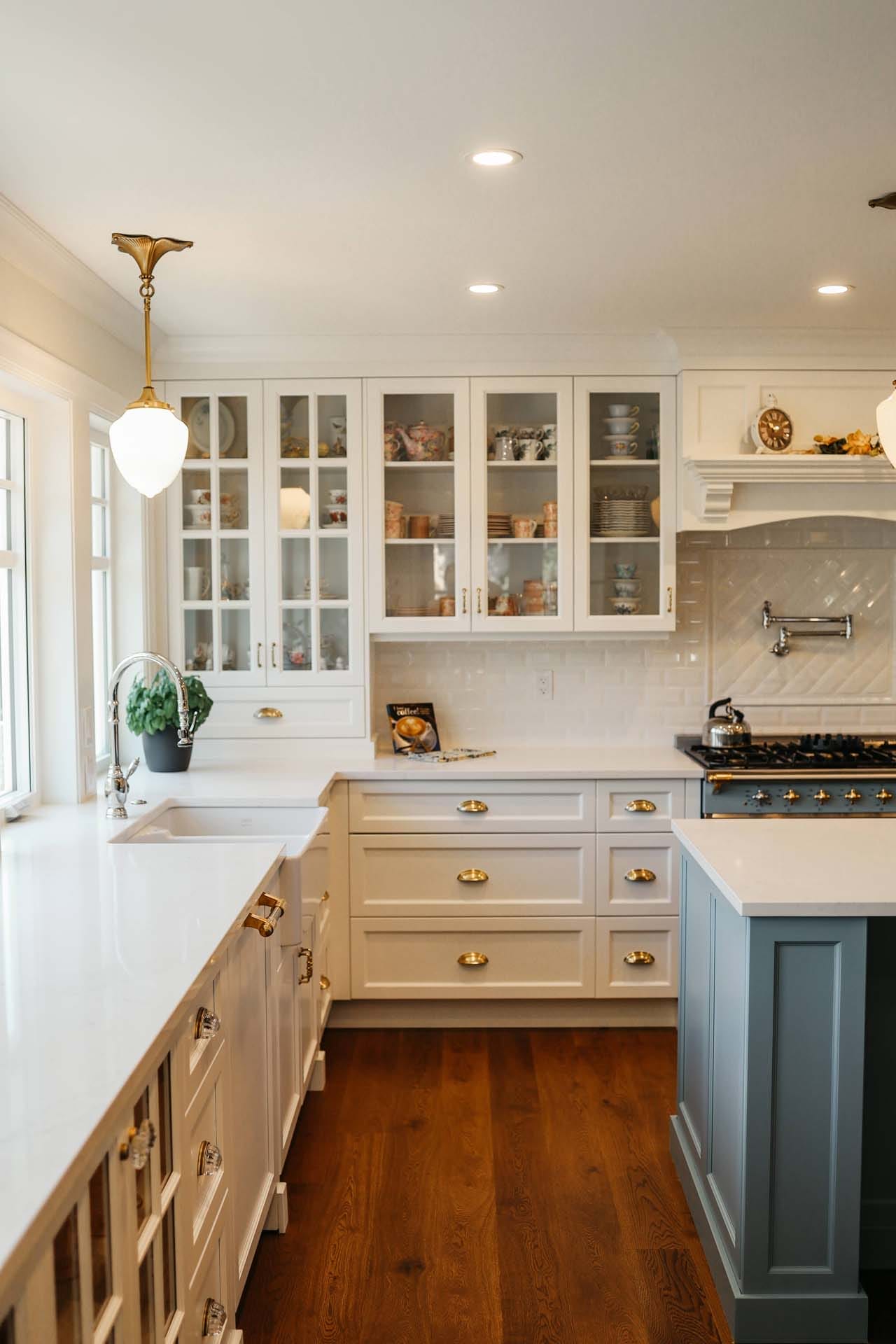 A modern kitchen with custom home design features, including white cabinets, glass-paneled cupboard doors, a marble countertop, and a green island. Wooden floors complement the style, while two pendant lights elegantly illuminate the island.