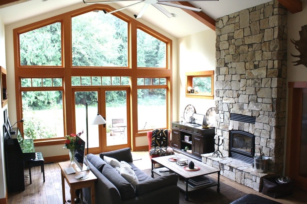 Spacious living room with large windows, crafted by expert home designers. Features a stone fireplace, sofa, coffee table, and a piano. Wood accents and greenery visible outside enhance the custom home design's bright and airy atmosphere.