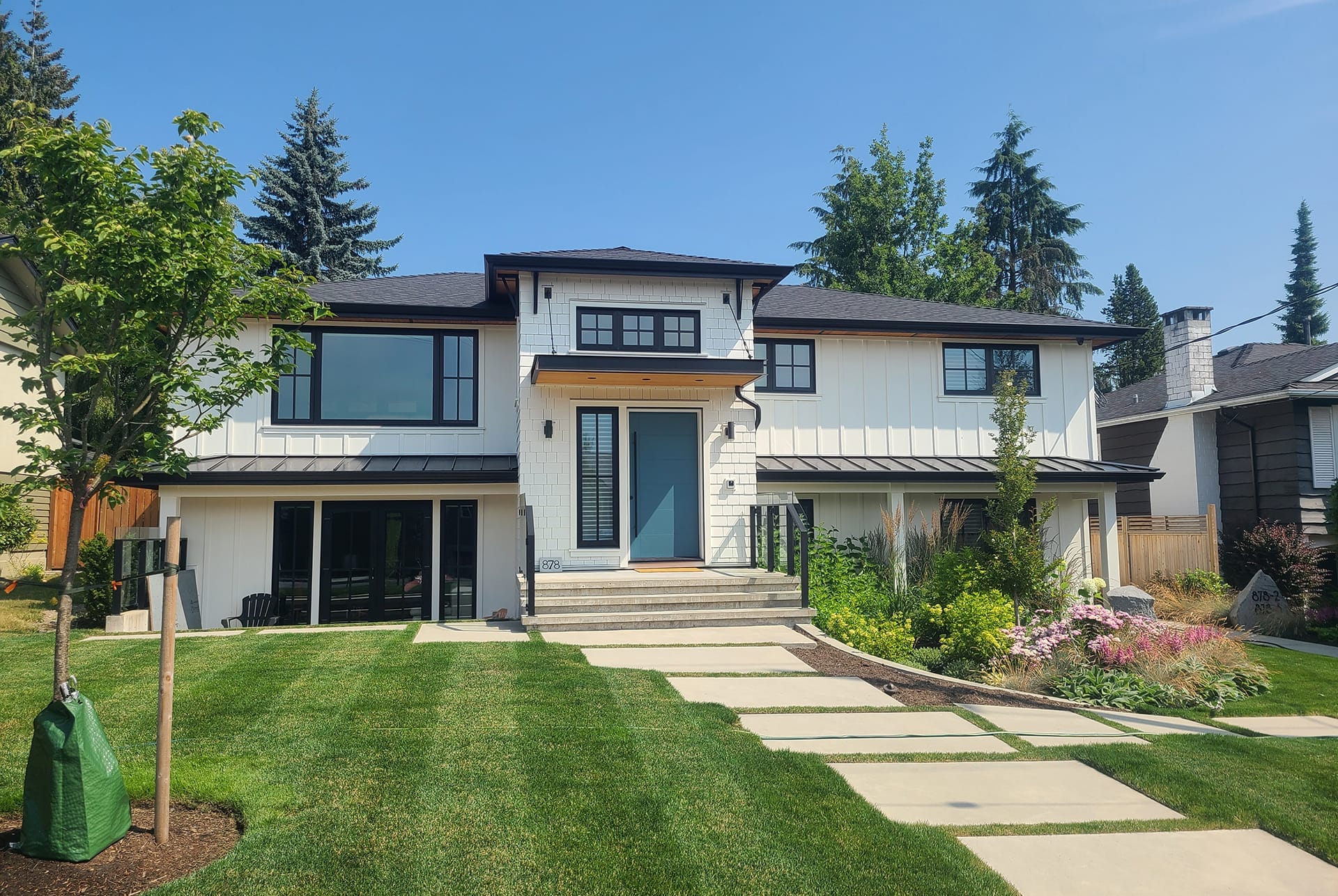 contemporary-home-renovation-project This two-story modern house features a custom home design with large windows, white siding, and dark trim. A manicured front lawn and a small tree on the left create a welcoming ambiance. Steps gracefully ascend to a central blue door, reflecting the expertise of skilled building designers.