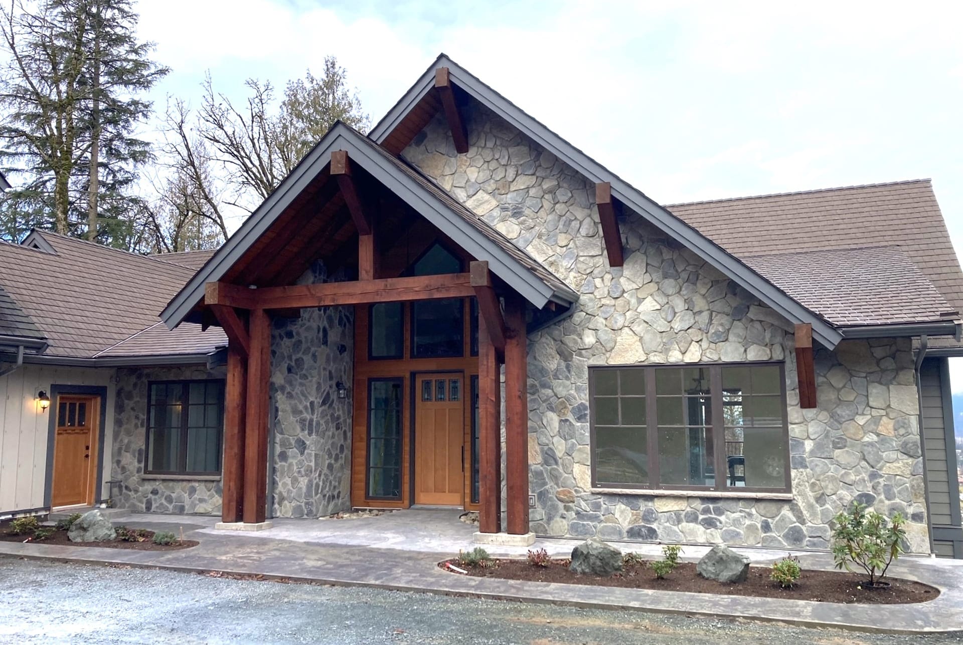 craftsman-renovation-project A stone house with a wooden door, large wooden beams, and a gabled roof crafted by building designers. The exterior boasts multiple windows, surrounded by a paved area and sparse landscaping.