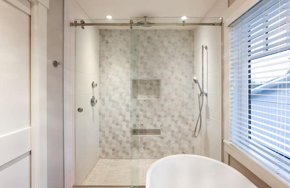 Modern bathroom with a glass-enclosed shower, white tub, and natural light from a window with blinds, showcasing the expertise of building designers in crafting custom home designs.