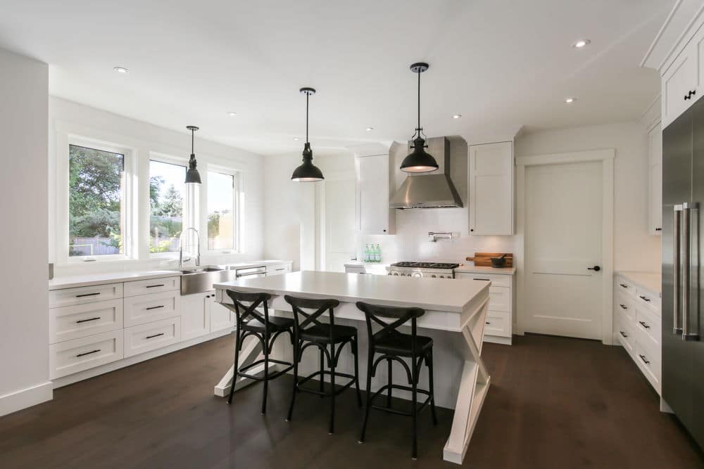 Modern kitchen with white cabinetry, a central island with three barstools, stainless steel appliances, and pendant lights. Large windows provide natural light in this custom home design curated by top building designers.