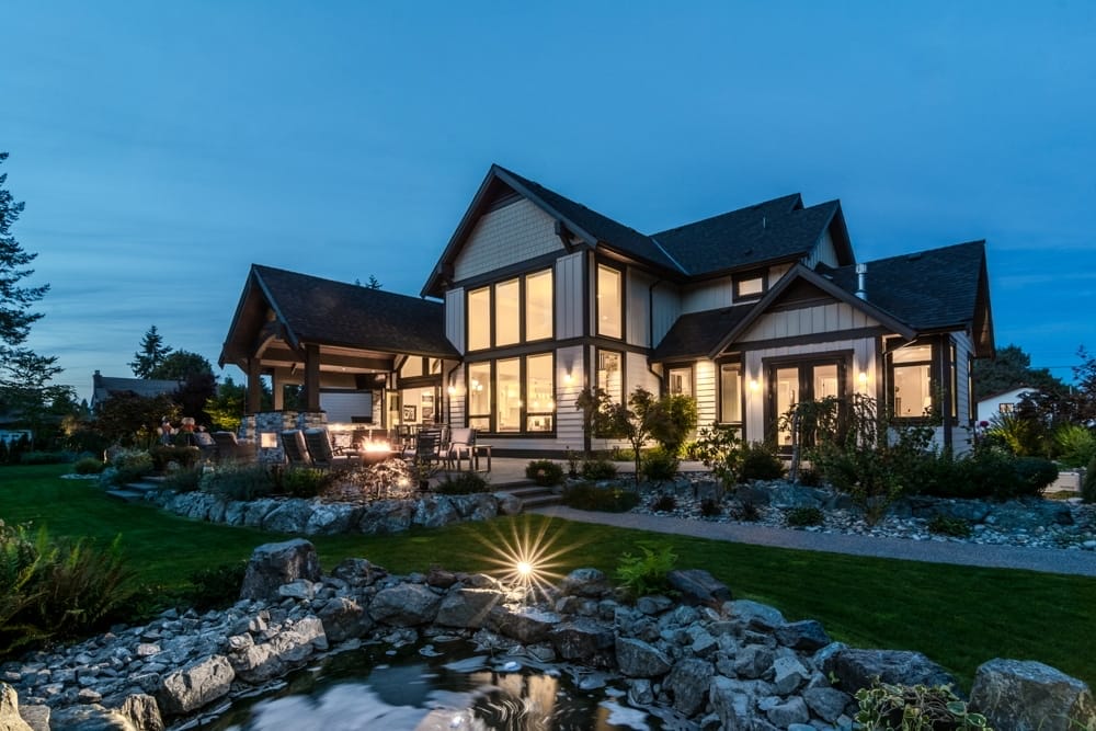 gables-galore-exterior-back-4 A modern house, crafted by home designers, boasts large windows lit from within at dusk. The landscaped yard features a pond with a water feature in the foreground.