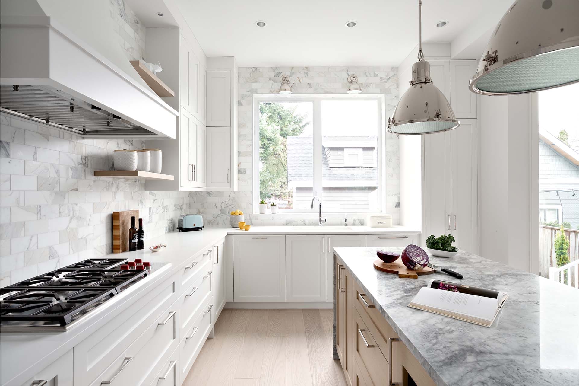 modern-home-interior-5 (5) Bright kitchen with white cabinets, marble countertops, and a gas stovetop, reflecting the expertise of home designers. An island centers the space where tableware and a cookbook rest. Two pendant lights hang above, while a window provides a view of the neighboring house.