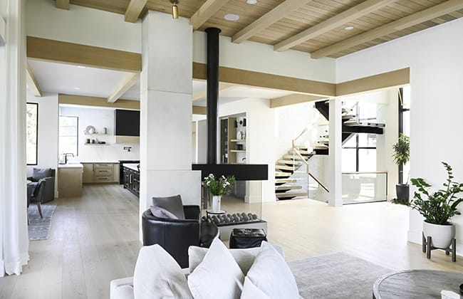 contemporary-home-main-floor This modern open-concept living area, crafted by skilled building designers, features wood beams, a suspended fireplace, a spiral staircase, and a kitchen in the background. Light wood flooring and potted plants accent the custom home design beautifully.