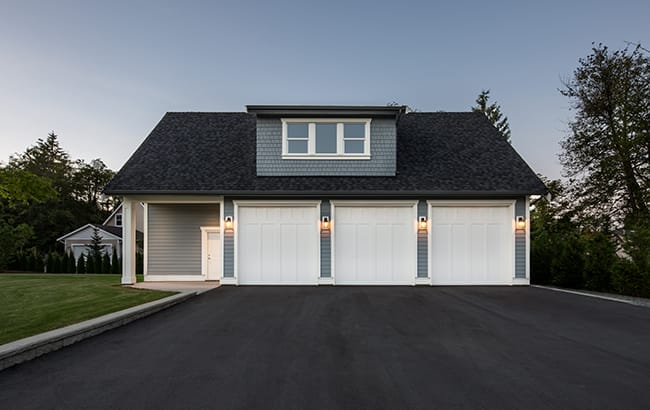 project3-traditional-carriage-house-design A large three-car garage with a gray and white exterior, expertly crafted by home designers, features a concrete driveway and is set against the backdrop of trees at dusk.