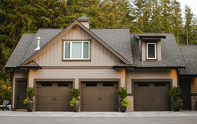 project5-carriage-house-design-example-2 A three-car garage with brown doors, gray siding, and a gabled roof showcases custom home design. Large planters with green plants adorn the front, while tall trees provide a scenic backdrop crafted by expert home designers.