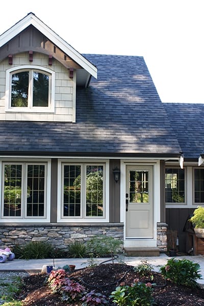 carriage-house-design-example-1 A charming custom home design features a gabled roof, stone facade, large windows, and a white door, all surrounded by a small garden.
