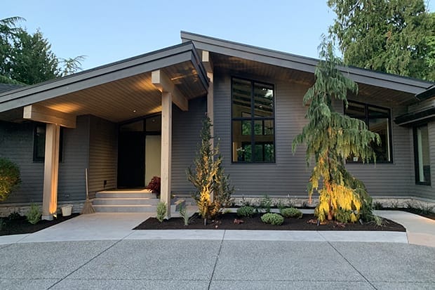 Modern single-story house with gray siding, large windows, and a sloped roof. Crafted by expert home designers, it features a small garden with coniferous trees and shrubs in front, reflecting a perfect blend of nature and custom home design.