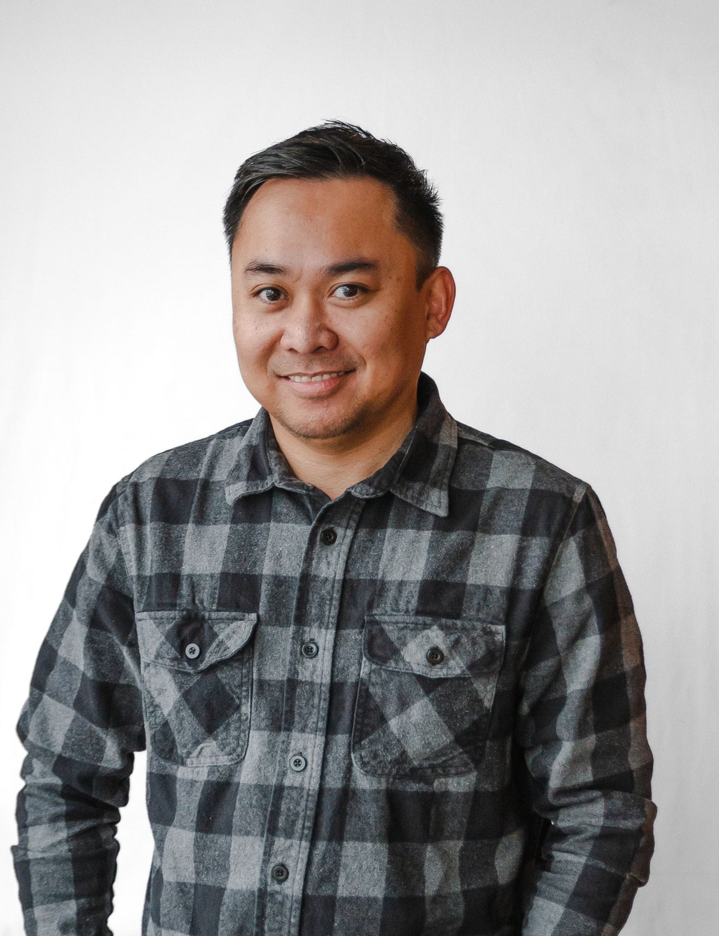 A person wearing a gray and black checkered shirt stands in front of a plain white background, smiling confidently like a seasoned home designer.
