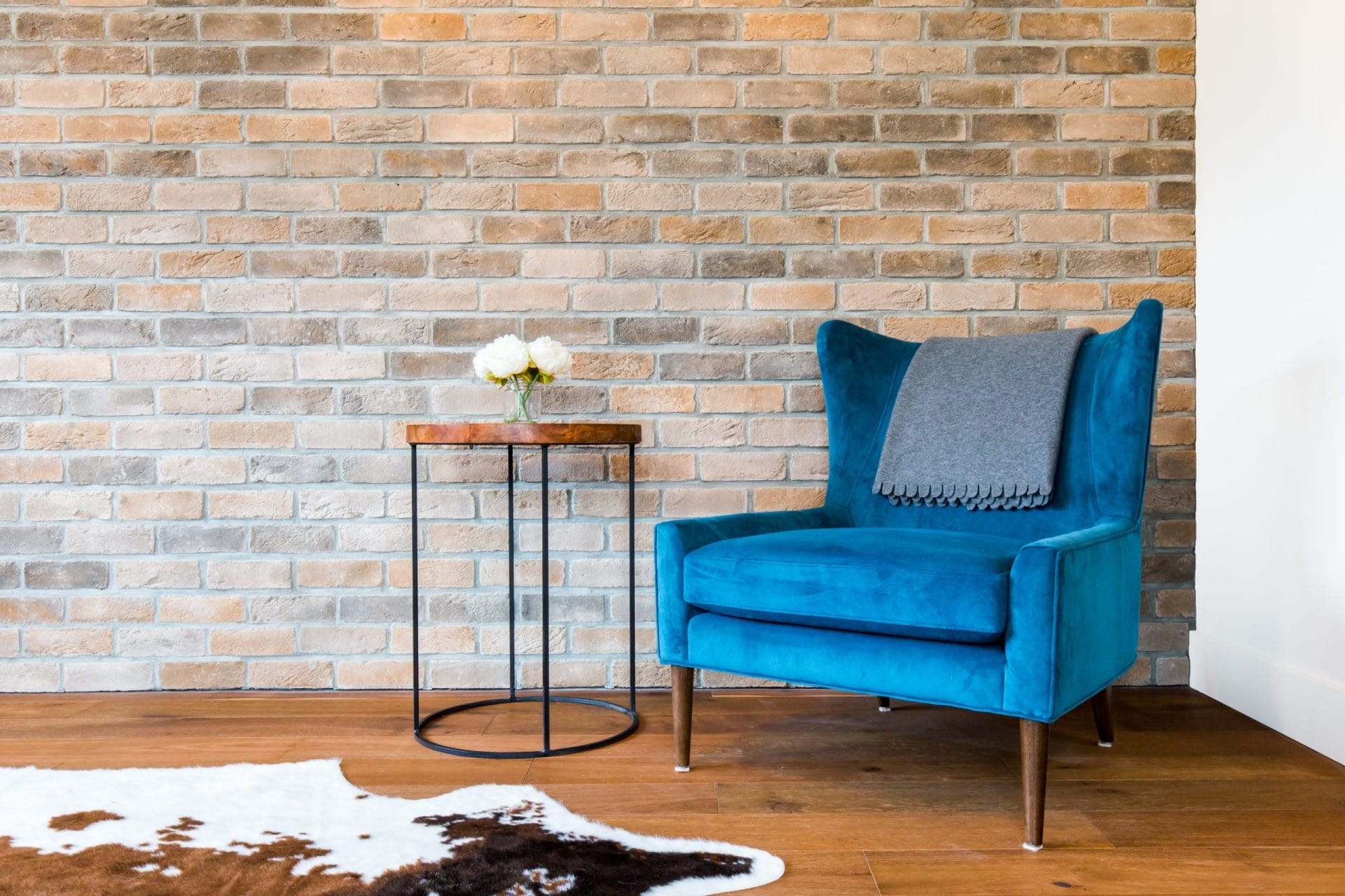 Blue armchair with a gray blanket beside a small round table holding white flowers, showcasing exquisite custom home design. A brick wall and cowhide rug complete this stylish setting.