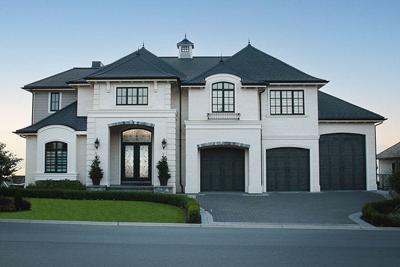 french-country-custom-home A custom home design showcases a large two-story house with a slate roof, three garages, and ornate front doors, surrounded by a manicured lawn and bushes.