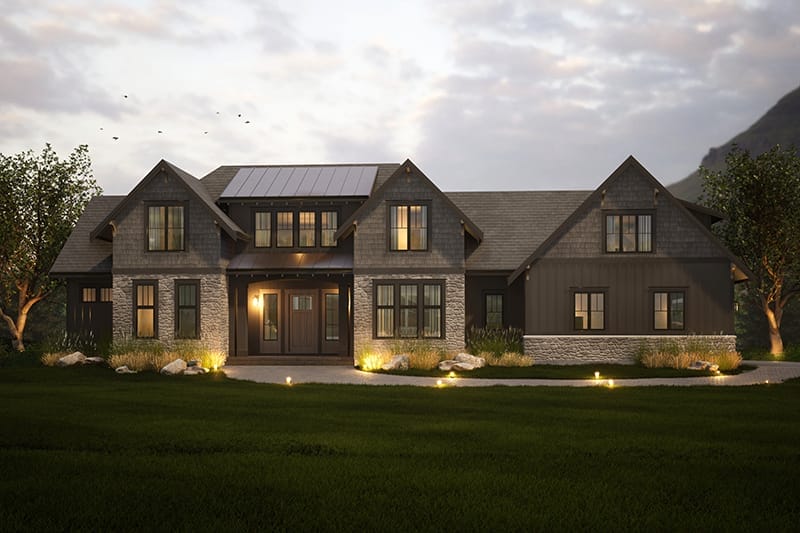 A modern rustic house exterior at dusk, thoughtfully crafted by home designers, features warm lights, multiple gabled roofs, and a harmonious mix of stone and wood siding. It's nestled amid lush greenery with majestic mountains standing serenely in the background.