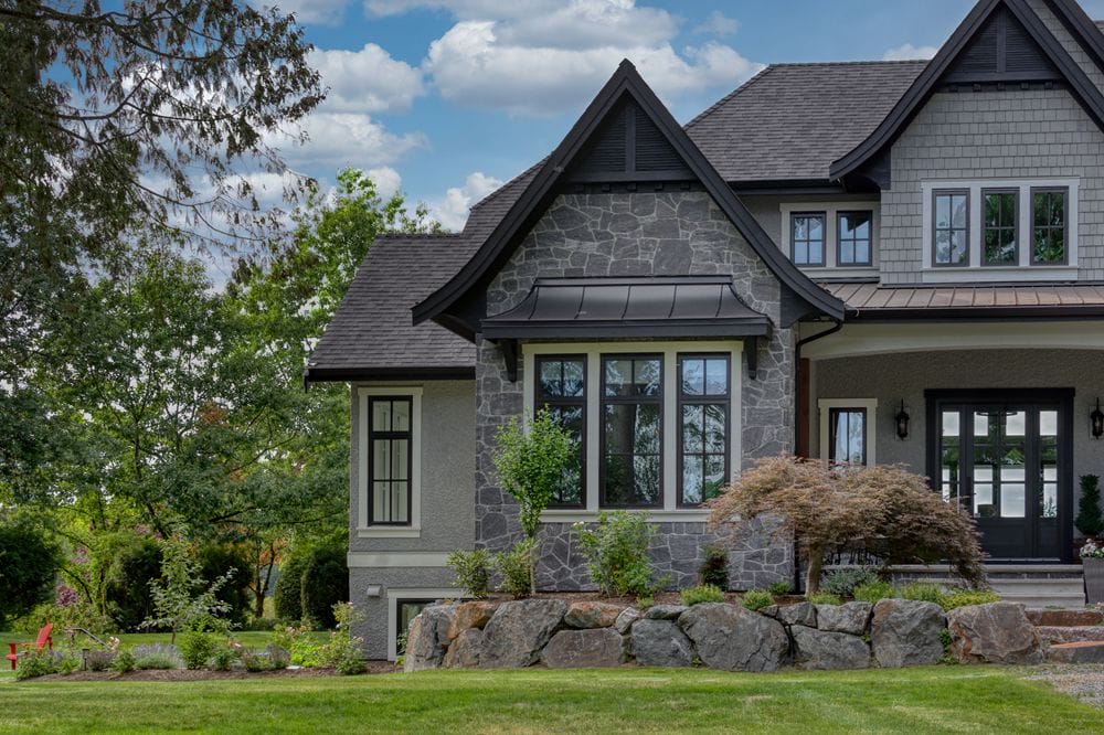 custom-craftsman-home-closeup A gray stone and stucco house with a black roof, nestled among trees and a well-maintained lawn, showcases the artistry of custom home design. The partly cloudy sky adds to the serene ambiance.