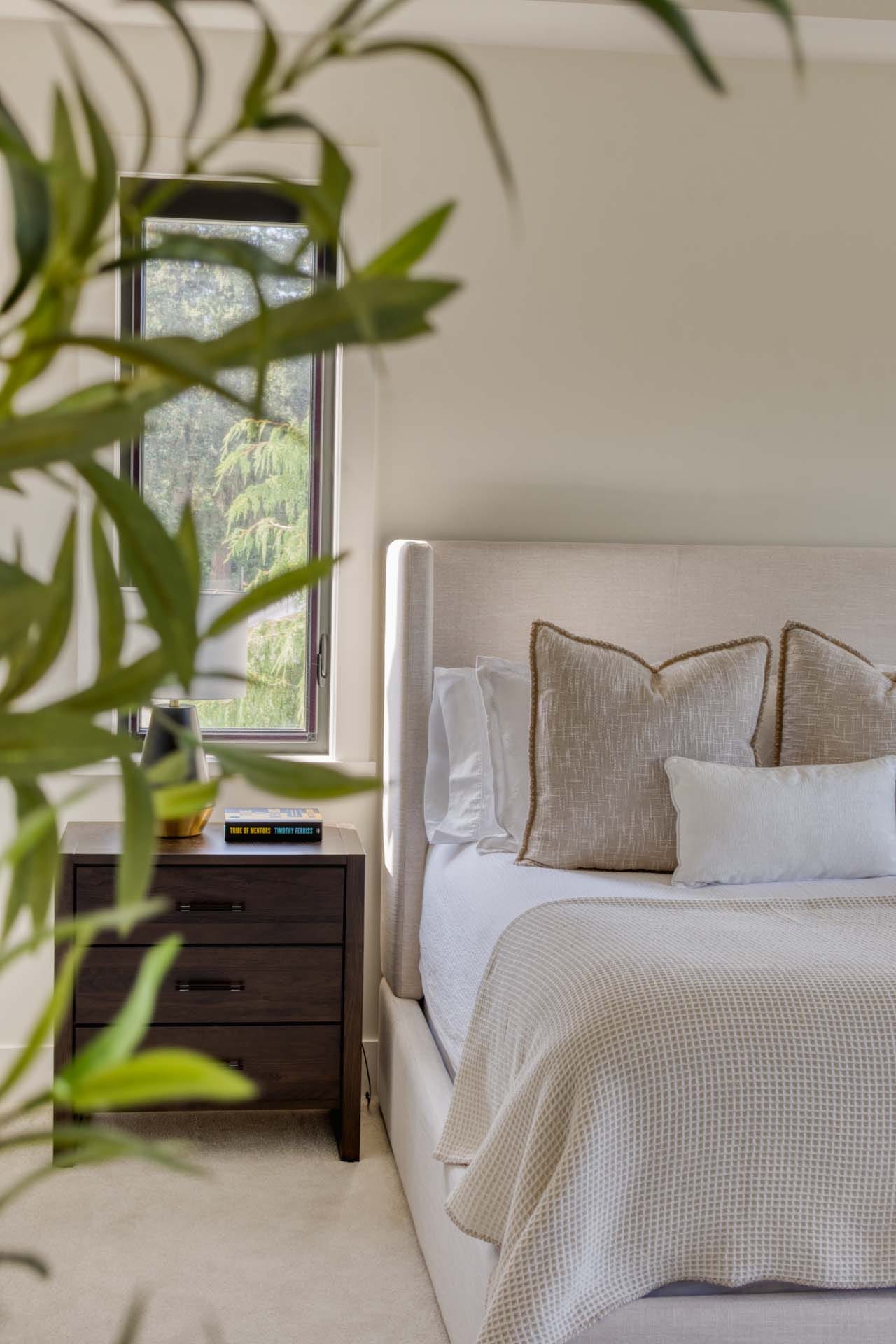 Neutral-toned bedroom designed by expert home designers, featuring a double bed, plush pillows, and a nightstand holding a book and vase. A leafy plant is partially visible in the foreground, adding a fresh touch to this custom home design.