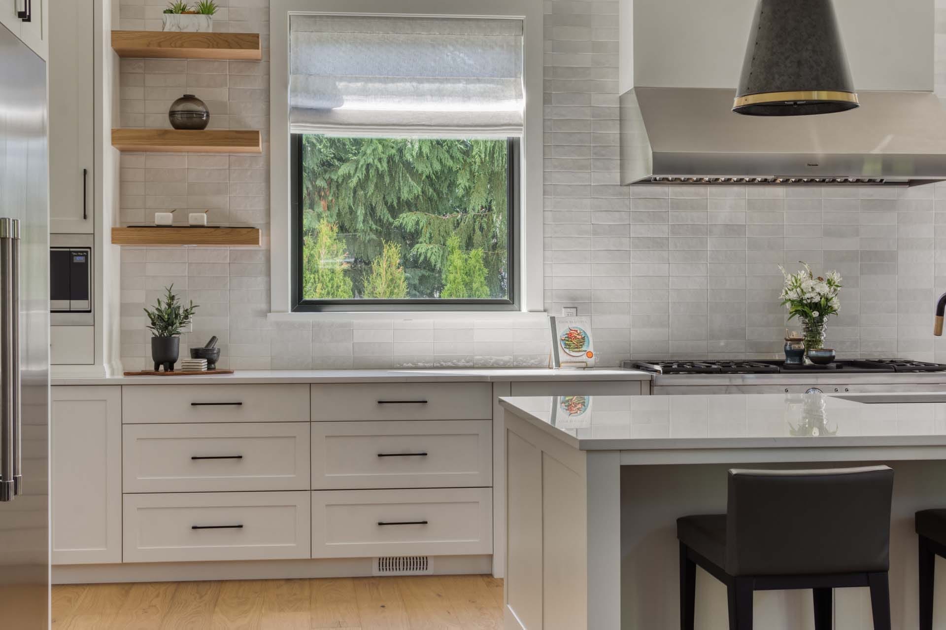 Modern kitchen by leading home designers, featuring white cabinetry, floating wooden shelves, a central island with two black chairs, and a window overlooking greenery.