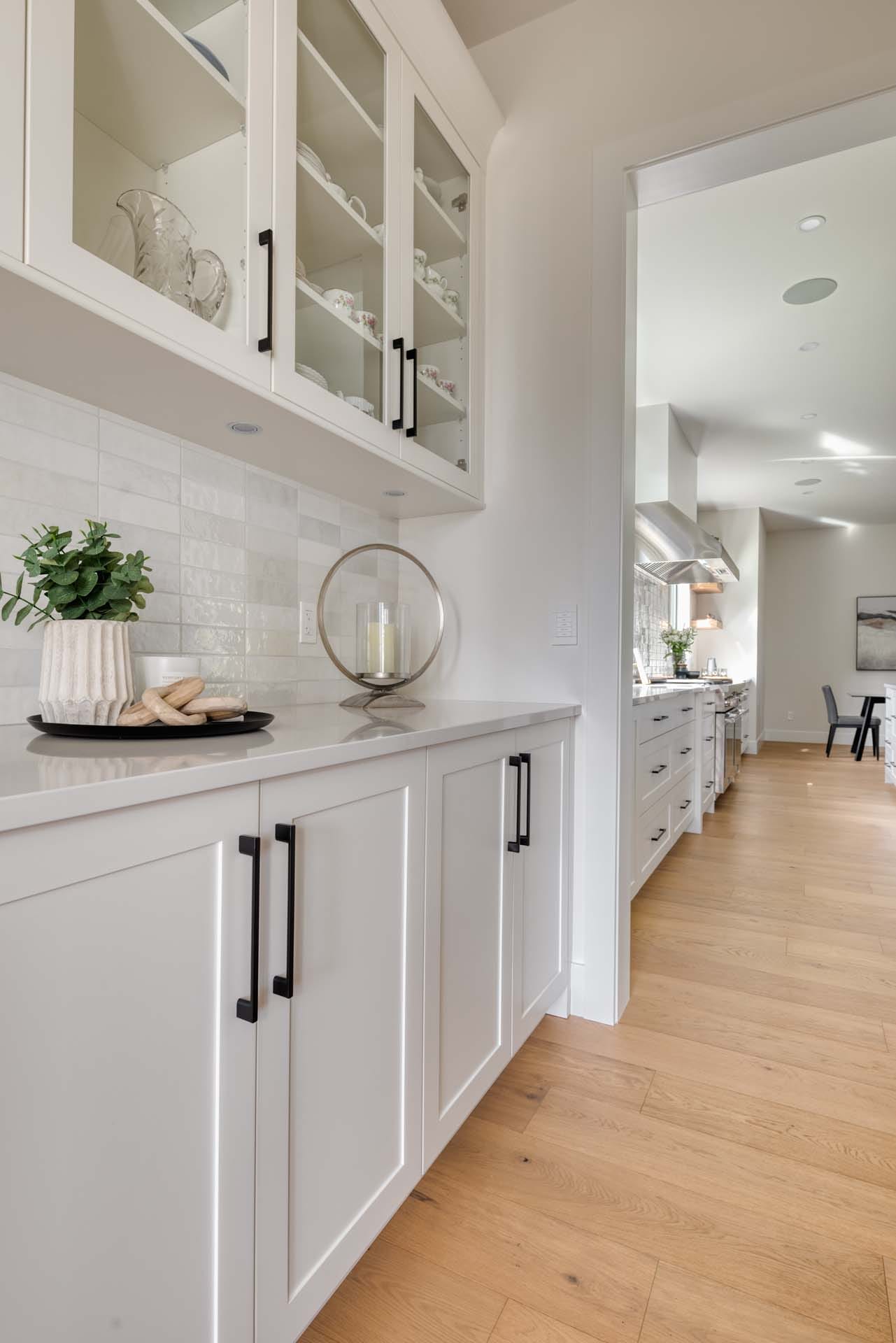This modern kitchen boasts white cabinets with glass doors and black handles, reflecting the artistry of custom home design. Wooden flooring seamlessly leads to a dining area, where a plant and tray with items add a touch of elegance crafted by expert home designers.