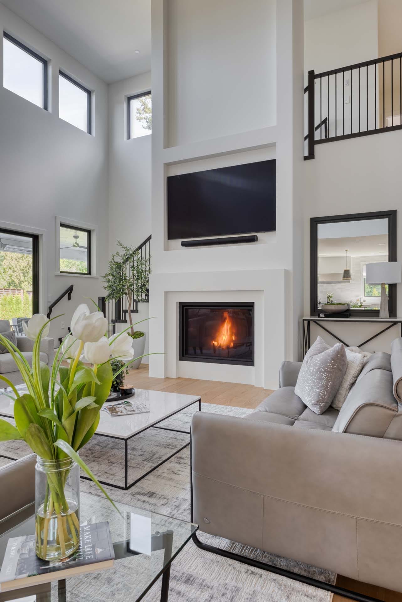 In this modern living room, artful elements by home designers feature a high ceiling and large windows. A sleek gray sofa faces a fireplace under a wall-mounted TV, while a glass vase with white flowers adorns the coffee table, embodying the essence of custom home design.
