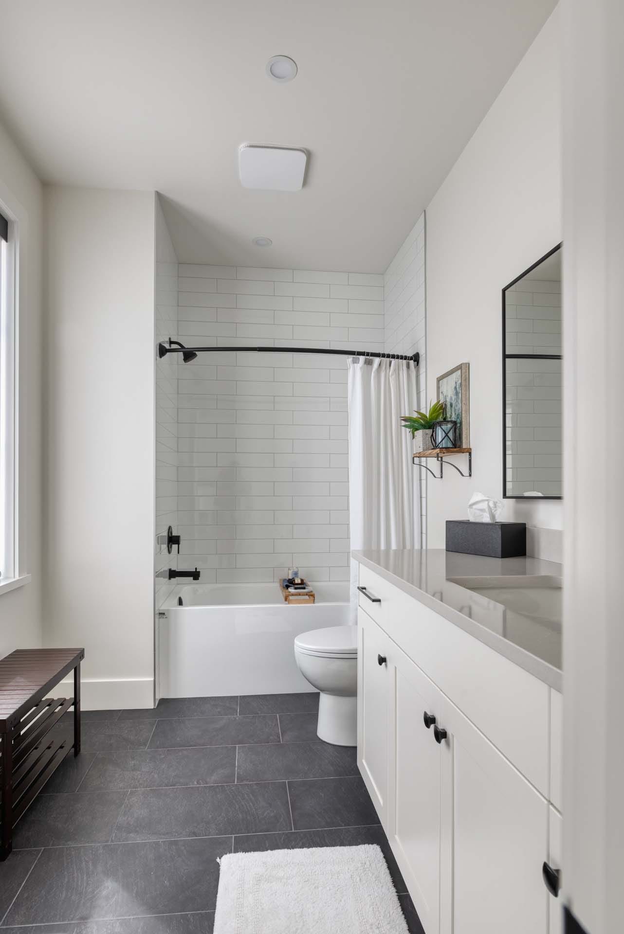Modern bathroom with a white bathtub and shower, black fixtures, a large mirror, and a vanity. White walls and a black-tile floor complement the minimalist decor, showcasing the expertise of building designers in custom home design.