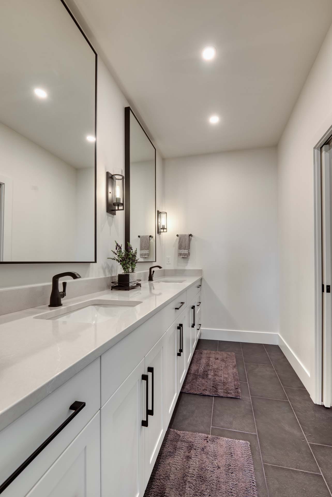 Modern bathroom crafted by top home designers, featuring a double sink vanity, large mirrors, and dark floor tiles. Wall lights illuminate the space above the sinks, while a plant and towels add charm to the custom home design on the countertop.