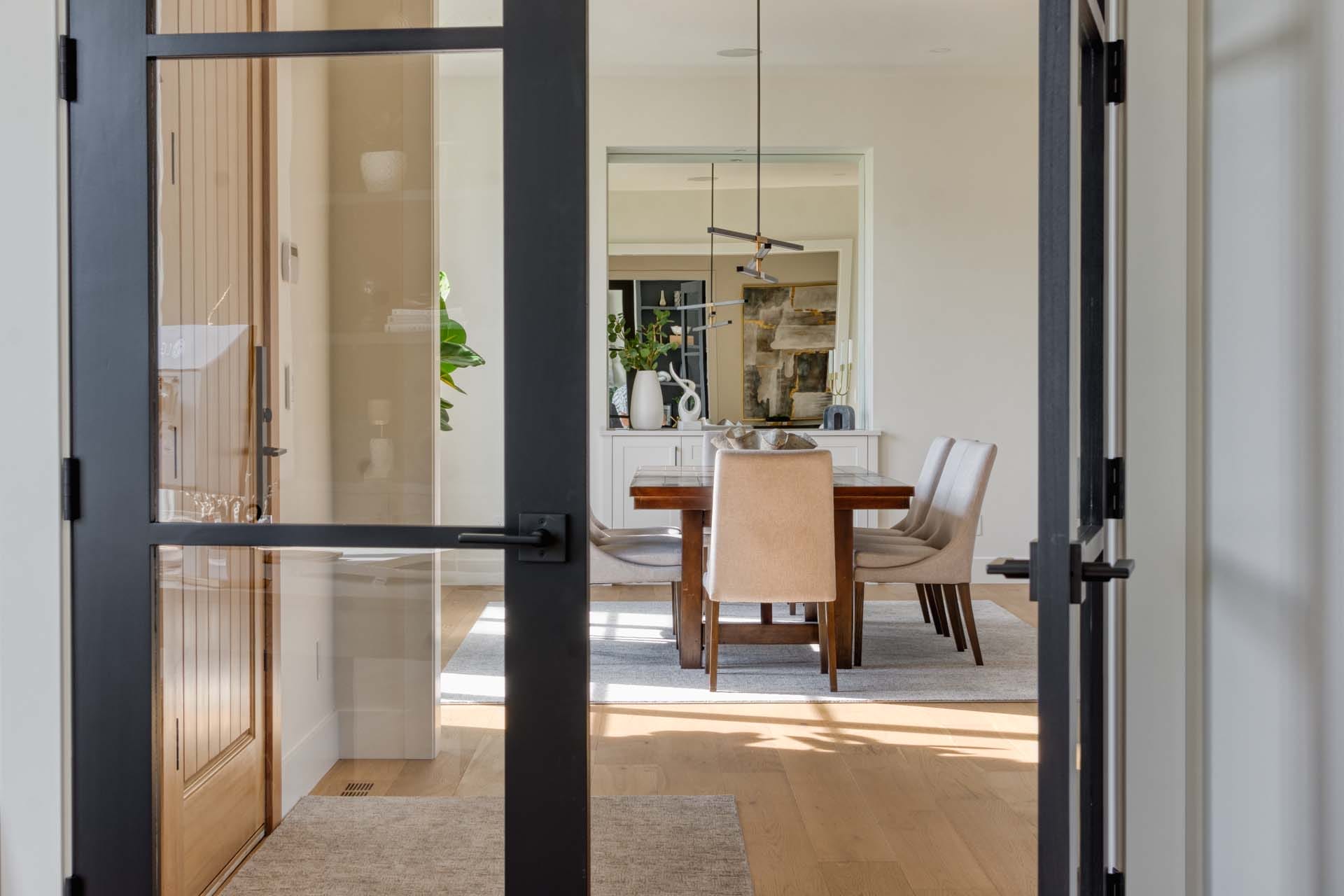 View through glass doors of a modern dining room, showcasing the touch of expert home designers with a wooden table, beige chairs, a light fixture, and a plant adding a natural accent.