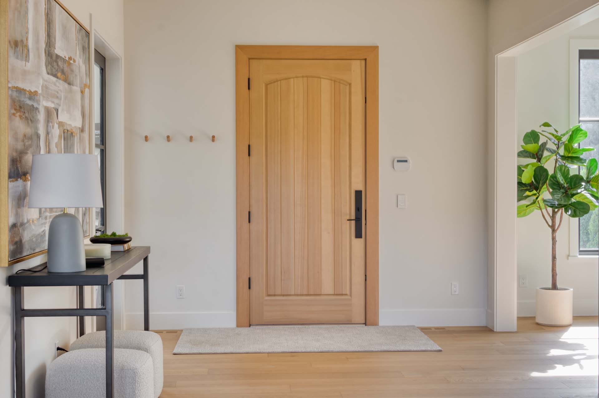 A minimalist entryway crafted by building designers features a wooden front door, light-colored walls, a side table with decor, a potted plant, and a small ottoman.