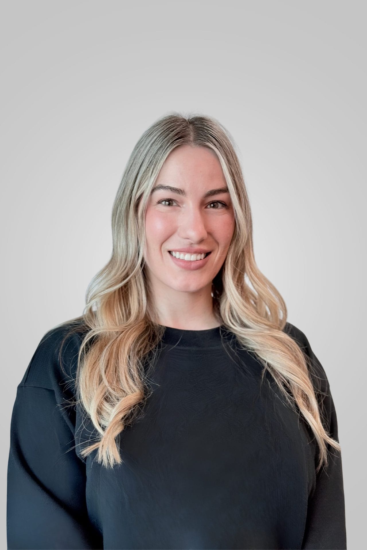 sam-pennington Person with long blonde hair wearing a black top stands against a plain gray background, smiling at the camera.