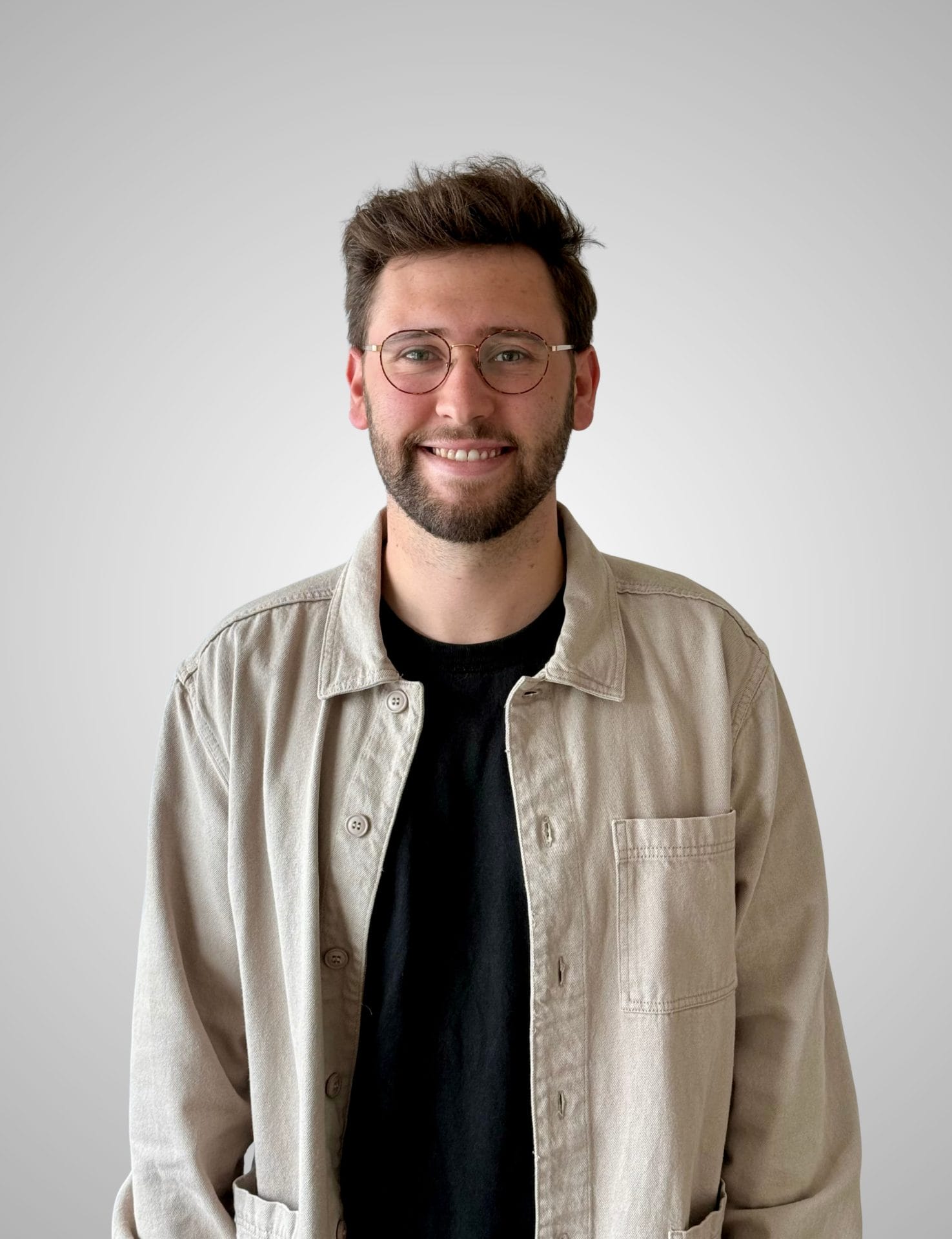 clayton-vanderveen-rev A man with short brown hair, glasses, and a beard is wearing a beige jacket over a black shirt, standing against a plain light gray background and smiling.