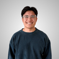 A person with short dark hair and glasses is smiling, wearing a dark ribbed sweater, standing against a plain light gray background.