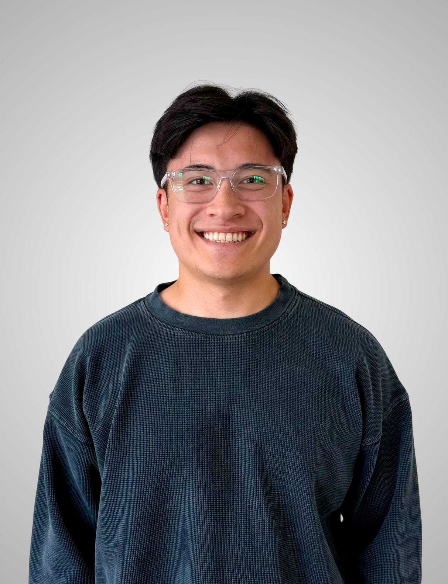 A person with short dark hair, glasses, and a dark sweatshirt is smiling and standing in front of a plain light gray background.